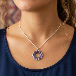 Puede incluir: Un collar de cadena de cuerda plateada con un colgante circular. El colgante está formado por un anillo de pequeñas cuentas cuadradas iridiscentes de color azul oscuro y morado, intercaladas con diminutas cuentas espaciadoras plateadas.