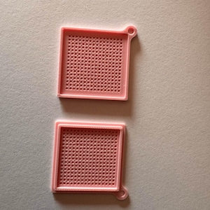 Puede incluir: Dos bandejas cuadradas de plástico rosa claro con una cuadrícula de pequeñas hendiduras circulares en la base. Cada bandeja presenta un borde elevado y una pequeña pestaña circular con un orificio en una esquina para colgar o sujetar.