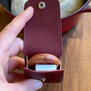 May include: A claret leather matchbox holder with a silver stud fastener, containing a wooden match striker and a box of matches. The holder is open, displaying the contents. A red pot and a loaf of bread are in the background.