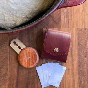 May include: A wooden, round, red-brown, shaving soap container with a silver razor blade and a burgundy leather case with a silver button. There are also four white envelopes on a wooden surface.