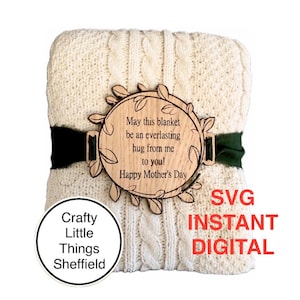 May include: A cream cable knit blanket with a wooden circular tag that reads "May this blanket be an everlasting hug from me to you! Happy Mother's Day." A dark green ribbon secures the tag. The words "SVG INSTANT DIGITAL" are in red.