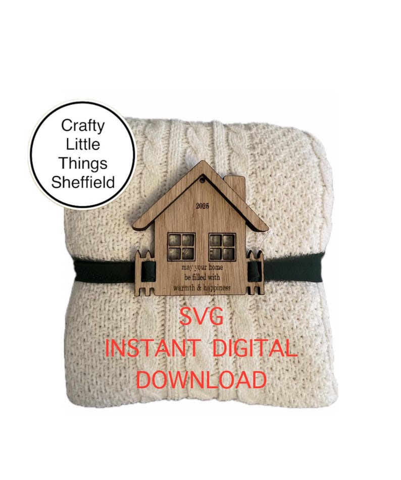 May include: A digital download SVG file of a house with the text "may your home be filled with warmth & happiness" and the year "2025". The house is on top of a cream-coloured knitted blanket with a black ribbon.