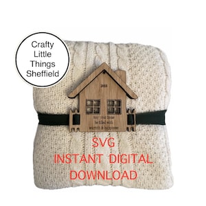 May include: A digital download SVG file of a house with the text "may your home be filled with warmth & happiness" and the year "2025". The house is on top of a cream-coloured knitted blanket with a black ribbon.