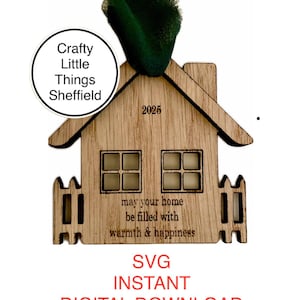 May include: A wooden house-shaped ornament with a white picket fence, windows, and the text "may your home be filled with warmth & happiness". The ornament is decorated with the year "2025" and the words "Crafty Little Things Sheffield".