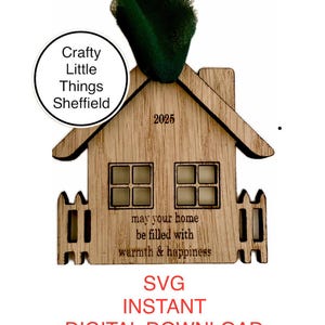 May include: A wooden house ornament with a white picket fence, windows, and the text "may your home be filled with warmth & happiness". The ornament is labelled "Crafty Little Things Sheffield" and "2025".