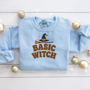 May include: A light blue crewneck sweatshirt featuring a graphic of a black witch hat and a broomstick above the text "BASIC WITCH" in a brown and black checkered pattern. The design includes small orange star accents around the hat.