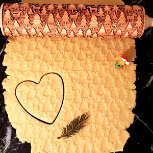 Puede incluir: Un rodillo de madera con un diseño de árbol de Navidad se utiliza para estirar la masa de galletas. La masa está estampada con el diseño del árbol. El rodillo tiene el texto "Carla Wood Art" en el mango.