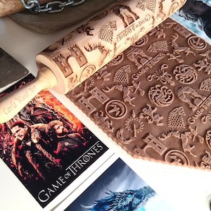 May include: A wooden rolling pin with a Game of Thrones theme. The rolling pin is engraved with various symbols and the words "Game of Thrones". The rolling pin is lying on a brown surface with a Game of Thrones poster in the background.