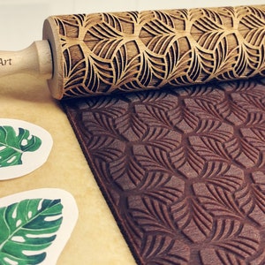 Puede incluir: Un rodillo de madera con un patrón de hojas talladas. El rodillo está sobre una superficie marrón con un patrón de hojas impreso en la superficie. El rodillo está grabado con el texto "Carla Wood Art".