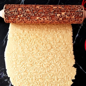 Puede incluir: Un rodillo de madera con un patrón de gatos está rodando sobre masa, creando un diseño en relieve. El rodillo tiene el texto "Carla Wood Art" en el mango. Un juguete de plumas azules y una madeja de lana roja también están en la imagen.
