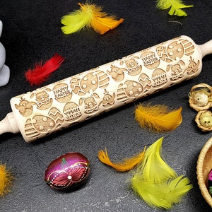 Peut inclure: Un rouleau à pâtisserie en bois gravé d'un motif d'œufs de Pâques, de poussins et des mots "Joyeuses Pâques". Le rouleau à pâtisserie est sur une surface noire avec des plumes jaunes et rouges, une figurine de lapin blanc et des œufs de Pâques décorés.