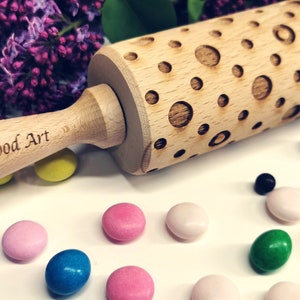May include: A wooden rolling pin with a carved pattern of circles. The rolling pin is engraved with the text "Carla Wood Art".