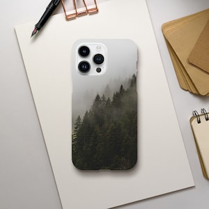 Könnte beinhalten: Eine weiße Smartphone-Hülle mit einer nebligen Waldszene. Das Bild zeigt einen Hügel mit immergrünen Bäumen, der teilweise von Nebel verdeckt ist. Die Hülle hat eine glänzende Oberfläche und ein Kameramodul mit drei Objektiven.