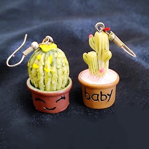 Könnte beinhalten: Ein Paar ausgefallene Ohrringe mit Miniatur-Kakteen in braunen Töpfen. Ein Ohrring hat einen runden Kaktus, der andere einen mehrstämmigen Kaktus. Die Töpfe haben lächelnde Gesichter und das Wort "baby".