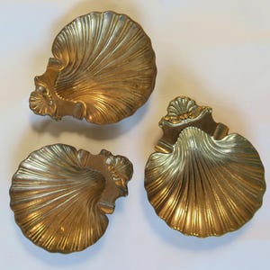 May include: Three antique brass seashell-shaped dishes. Each dish has a detailed, ridged design, with a scalloped edge and a small decorative element at the top. The dishes are arranged on a white surface.