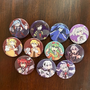 Op de afbeelding: Een verzameling van elf ronde buttons met kleurrijke illustraties van personages in anime-stijl. Elke button toont een ander personage met unieke kapsels, kleding en uitdrukkingen. De buttons zijn gerangschikt op een donker houten oppervlak.