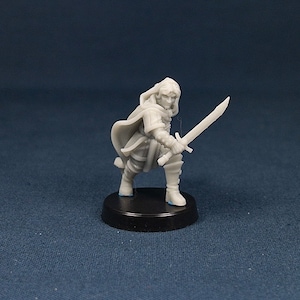 May include: A gray plastic miniature figurine of a person wearing a hooded cloak and holding a sword. The figurine is standing on a black base.