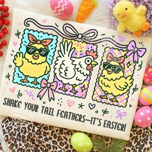 May include: A cream-colored tote bag featuring three cartoon chickens and the text "SHAKE YOUR TAIL FEATHERS - IT'S EASTER!". The chickens are in colorful frames with bows and sunglasses. Easter eggs and a chick are in the background.