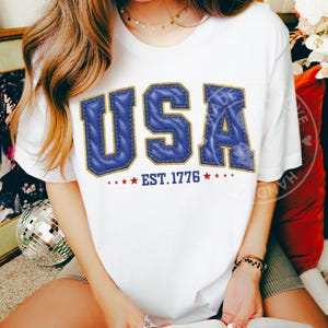 Puede incluir: Camiseta blanca con la palabra "USA" en letras azules acolchadas con ribete dorado. Debajo, estrellas rojas y el texto "EST. 1776". La camiseta es de cuello redondo.