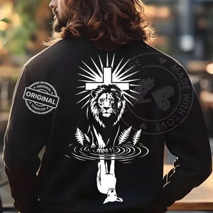 Puede incluir: Chaqueta negra con un diseño gráfico blanco de un león con una cruz encima. El león se refleja en el agua, con un cordero debajo. La palabra "ORIGINAL" está impresa en la manga izquierda.