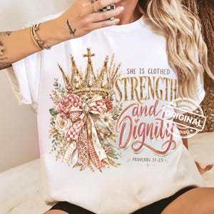 May include: A white t-shirt featuring a graphic print of a gold crown adorned with pink and white flowers, leopard print ribbons, and the text "SHE IS CLOTHED STRENGTH and Dignity PROVERBS 31:25" in gold and pink script lettering.