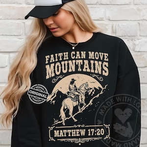 Könnte beinhalten: Schwarzes Sweatshirt mit dem Text "FAITH CAN MOVE MOUNTAINS" im Vintage-Stil. Das Design beinhaltet eine Illustration einer Person, die vor Bergen reitet, mit dem Bibelvers "MATTHEW 17:20" darunter. Das Sweatshirt hat einen "ORIGINAL" Stempel.