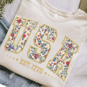 May include: Cream-colored t-shirt with the word "USA" in floral print letters outlined in gold glitter. The text "EST. 1776" is below the letters, with gold stars. The shirt is folded on a denim surface.