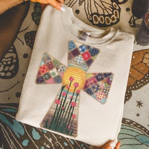 May include: Cream-colored sweatshirt featuring a cross design with a patchwork pattern in various colors. The cross has a yellow sun in the center, pink flowers, and green grass. The sweatshirt is folded and held by hands.