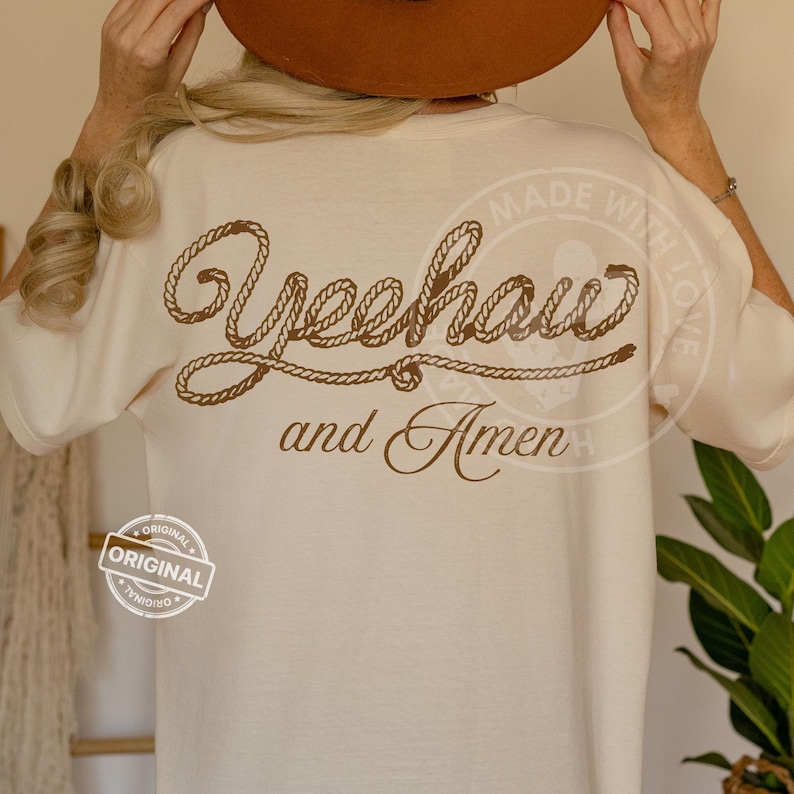 Peut inclure: T-shirt cr&egrave;me avec l'inscription "Yeehaw and Amen" en lettres marron fa&ccedil;on corde. Le t-shirt a une coupe d&eacute;contract&eacute;e et un col rond. Un petit tampon "Original" est visible.