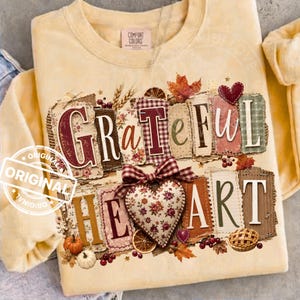 Puede incluir: Una sudadera de cuello redondo color amarillo pálido con un estampado gráfico que incluye el texto "GRATEFUL HEART" en estilo patchwork. El diseño presenta un corazón floral, hojas de otoño, espigas de trigo, una calabaza y una tarta.