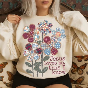 Puede incluir: Sudadera color crema con un diseño floral. Las flores tienen pétalos de estilo patchwork en varios patrones y colores. El texto "Jesus loves me, this I know" está impreso en marrón. Manga larga.