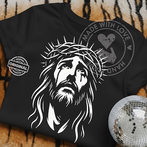 May include: Black t-shirt featuring a white graphic of Jesus wearing a crown of thorns. The design includes a detailed depiction of his face, with tears and long hair. The shirt also has the words "ORIGINAL" and "MADE WITH LOVE HAND" printed on it.
