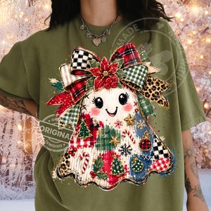 May include: An olive green t-shirt featuring a central graphic of a ghost decorated with Christmas elements. The ghost wears a large bow with plaid, leopard, and checkered patterns, adorned with a poinsettia, candy canes, trees, and ornaments.