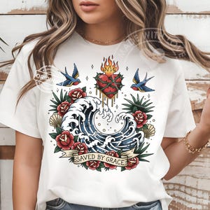 May include: White t-shirt featuring a tattoo-style design. The graphic includes a heart with flames, a wave, roses, swallows, and the text "SAVED BY GRACE" on a banner. The design is in red, blue, and green.