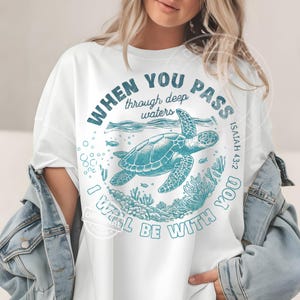 May include: White t-shirt featuring a teal sea turtle graphic. The text reads "WHEN YOU PASS through deep waters, I WILL BE WITH YOU" with "ISAIAH 43:2" below. The design is circular, depicting a turtle in the ocean.