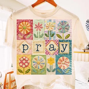 Könnte beinhalten: Cremefarbenes T-Shirt mit einem farbenfrohen Blumen-Patchwork-Design. Das Design umfasst verschiedene Blumen und das Wort "PRAY" in einer gestickten Schriftart. Das Shirt hängt an einem Holzbügel.