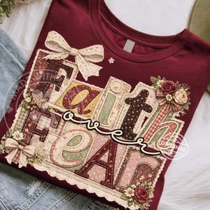 May include: Burgundy t-shirt with a patchwork design that reads "Faith over Fear." The design features floral patterns, lace, and bows. The letters are in various colors and textures, creating a vintage, handcrafted aesthetic.