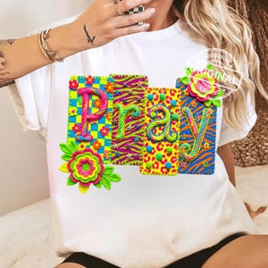 May include: White t-shirt with the word "PRAY" in colorful, 3D letters. Each letter has a unique pattern, including zebra stripes, leopard print, and floral designs. The letters are adorned with decorative flowers.