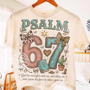 Könnte beinhalten: Cremefarbenes T-Shirt mit dem Text "PSALM 67" in Türkis und Rosa. Das Design umfasst einen Stern, Schleifen, ein Herz, eine Bibel, einen Kaktus und einen Cowboy-Stiefel. Das Shirt trägt auch das Zitat "Gott sei uns gnädig und segne uns; und lasse sein Angesicht leuchten über uns."