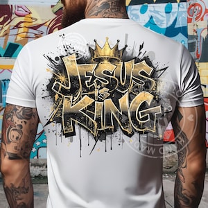 May include: White t-shirt with a gold and black graffiti-style design that reads "JESUS is KING". The design includes a gold crown and paint splatter effects. The shirt is a crew neck.