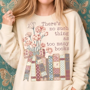May include: Cream-colored sweatshirt with a floral and book design. The graphic features a bouquet of pink flowers, a patchwork bow, and a stack of books. Text reads "There's no such thing as too many books."
