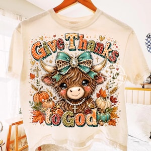 May include: A cream-colored t-shirt featuring a graphic of a brown highland cow with a leopard print bow, wearing a pearl necklace with a cross pendant. The text reads Give Thanks to God, surrounded by pumpkins, autumn leaves, and crosses.