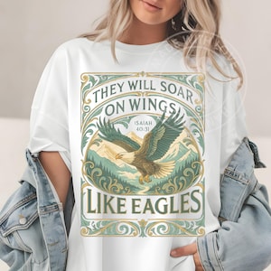 May include: White t-shirt featuring a detailed graphic of an eagle soaring over a mountain landscape. The design includes the text "THEY WILL SOAR ON WINGS LIKE EAGLES" and the Bible verse Isaiah 40:31, in a decorative frame.