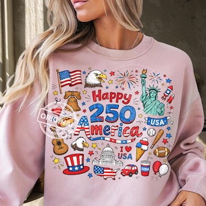 May include: A light pink sweatshirt with a colorful graphic celebrating America's 250th anniversary. The design includes the American flag, an eagle, the Statue of Liberty, fireworks, and other patriotic symbols.
