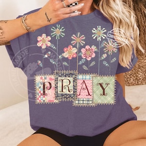 May include: A purple t-shirt with a floral design and the word "PRAY" in patchwork letters. The flowers are in various colors, including pink, blue, and green. The shirt has short sleeves and a relaxed fit.