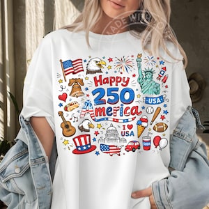 May include: White t-shirt with a colorful graphic celebrating America's 250th anniversary. The design includes an American flag, an eagle, the Statue of Liberty, fireworks, and other patriotic symbols.