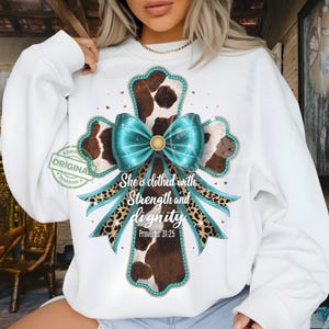 May include: White sweatshirt featuring a cross design with a cowhide pattern, teal bow, and leopard print accents. The text "She is clothed with Strength and dignity" is displayed below the cross. The design includes the Bible verse Proverbs 31:25.