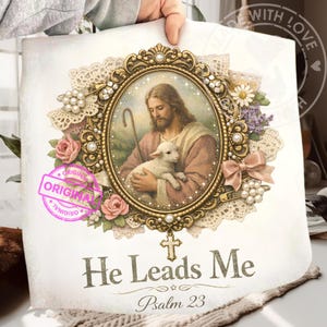 May include: A religious-themed print featuring an oval portrait of Jesus holding a lamb, framed in gold with floral and lace accents. The text "He Leads Me, Psalm 23" is displayed below the portrait. The print is held by a person.