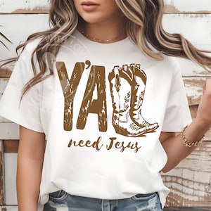 Puede incluir: Camiseta blanca con un gráfico marrón que dice "Y'all need Jesus" y presenta un par de botas de vaquero. El texto y las botas están en un estilo rústico y desgastado. La camiseta es una prenda informal.