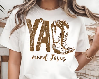 Könnte beinhalten: Weißes T-Shirt mit einem braunen Aufdruck, der "Y'all need Jesus" lautet und ein Paar Cowboy-Stiefel zeigt. Der Text und die Stiefel sind in einem rustikalen, verwitterten Stil gehalten. Das Shirt ist ein lässiges Kleidungsstück.